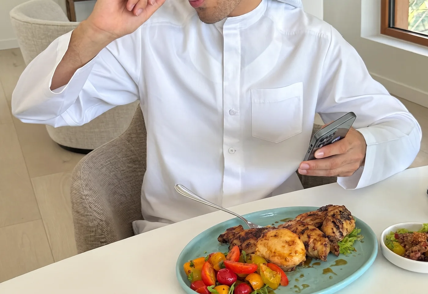 Man in white thobe sitting at a home dining table, logging his meal on his phone after eating